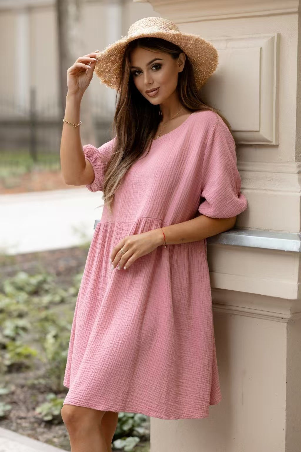 A woman wearing a pink textured short-sleeve dress and a straw sun hat, leaning against a cream-colored architectural column outdoors.