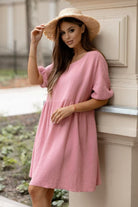 A woman wearing a pink textured short-sleeve dress and a straw sun hat, leaning against a cream-colored architectural column outdoors.