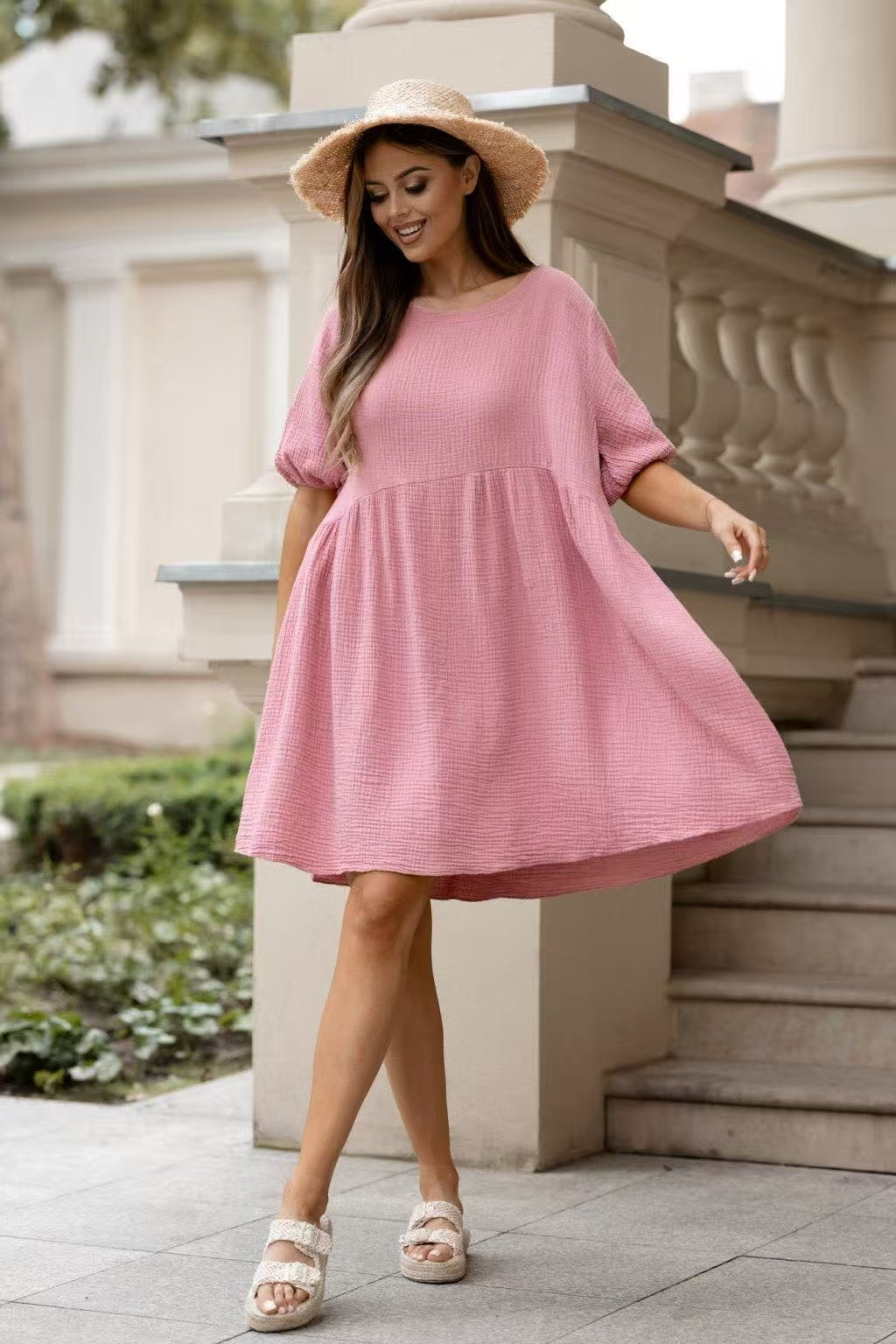 A woman wearing a pink textured short-sleeve dress with a gathered waist, styled with a straw hat and beige platform sandals, standing outdoors in front of a stone balustrade.