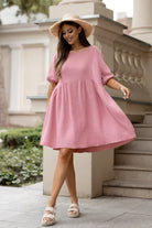 A woman wearing a pink textured short-sleeve dress with a gathered waist, styled with a straw hat and beige platform sandals, standing outdoors in front of a stone balustrade.