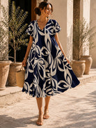 Woman wearing a blue and white patterned dress standing outdoors with plants and columns in the background.