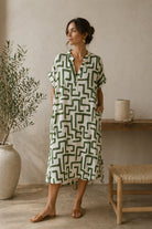 Woman wearing a green and white patterned dress standing in a room with a plant and wooden stool.