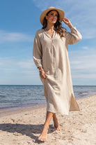 A woman stands on a sandy beach wearing a long-sleeve, midi-length beige textured dress with a V-neck and a wide-brimmed straw hat, with the ocean and blue sky in the background.