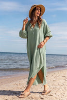 A woman wearing a light sage green waffle-knit maxi dress and a wide-brim straw hat walks on a sandy beach with the ocean and blue sky in the background.