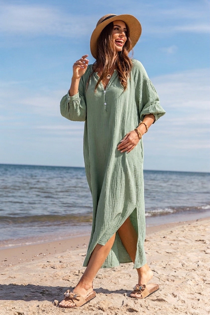 A woman wearing a light sage green waffle-knit maxi dress and a wide-brim straw hat walks on a sandy beach with the ocean and blue sky in the background.