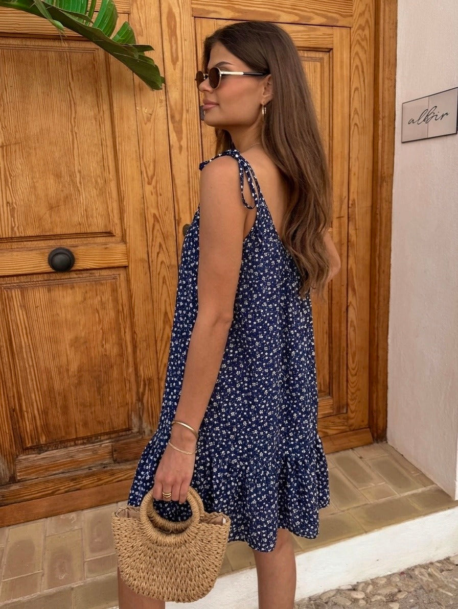 A woman wearing a navy blue sleeveless dress with a white floral print, standing in front of a rustic wooden door and holding a woven straw handbag.