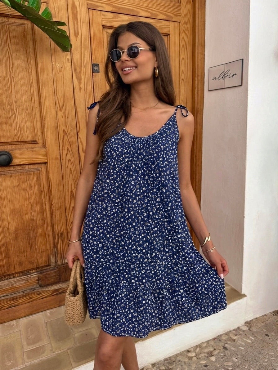 A woman wearing a navy blue sleeveless mini dress with a small white floral print, standing in front of a rustic wooden door and holding a woven straw bag.