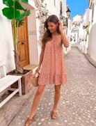 A woman wearing a light pink sleeveless dress with a small red and green floral print, standing on a cobblestone street in a white-walled village setting.