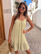 A woman wearing a light yellow sleeveless dress with a small white floral print, standing on a cobblestone street in a sunny, white-walled village setting.