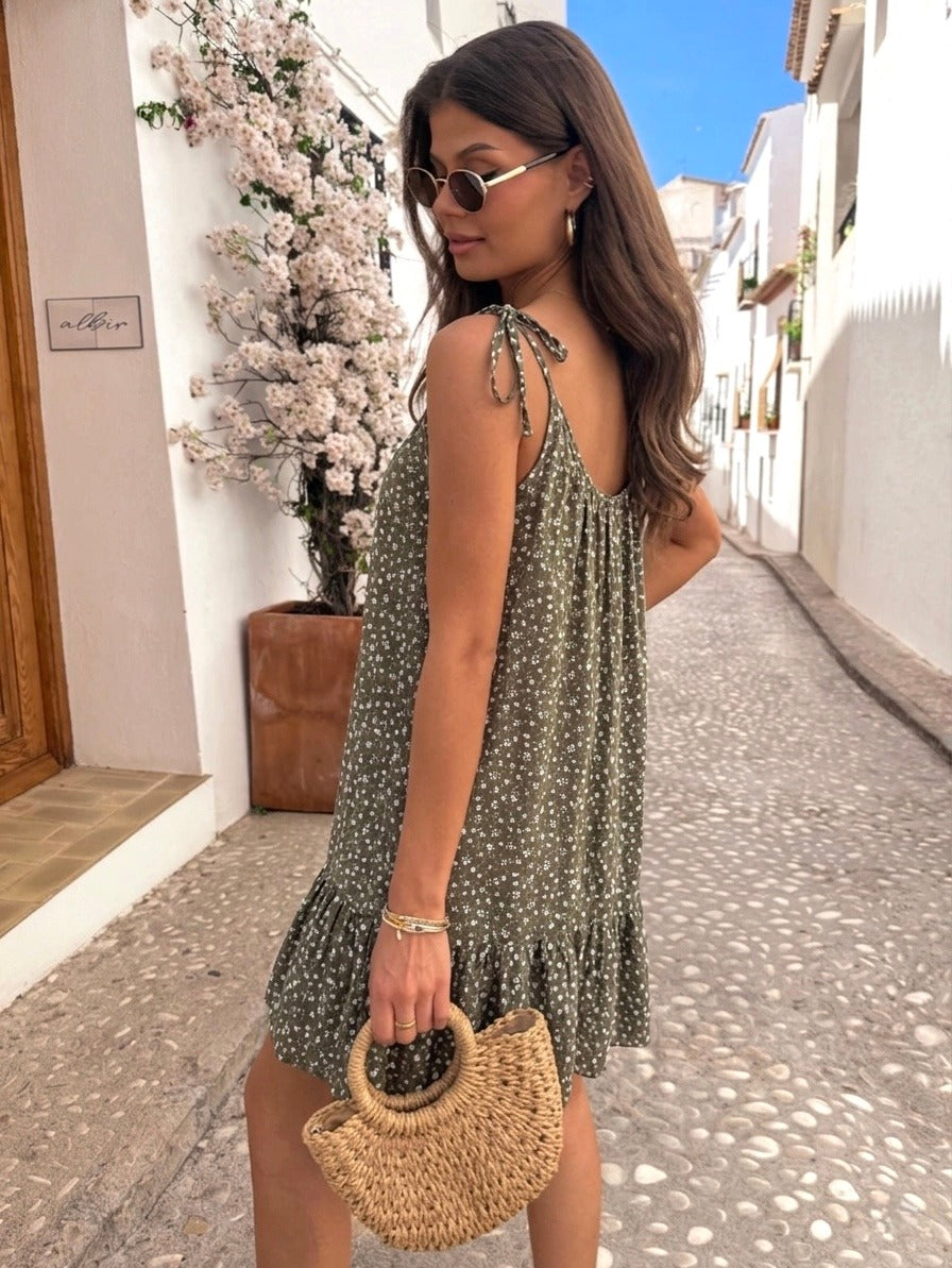 A woman wearing a sleeveless olive green and white floral print dress stands on a cobblestone street in a sunny, white-walled village setting, holding a woven straw handbag.