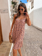 A woman wearing a sleeveless pink floral midi dress with a ruffled hem, standing in a sunny, narrow white-walled street. She is holding a woven straw clutch and wearing round sunglasses.