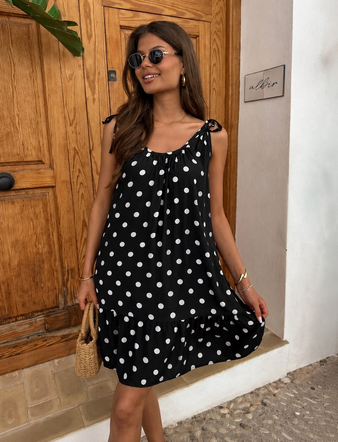 A woman wearing a black sleeveless dress with white polka dots and tie-shoulder straps, standing in front of a wooden door and white wall.