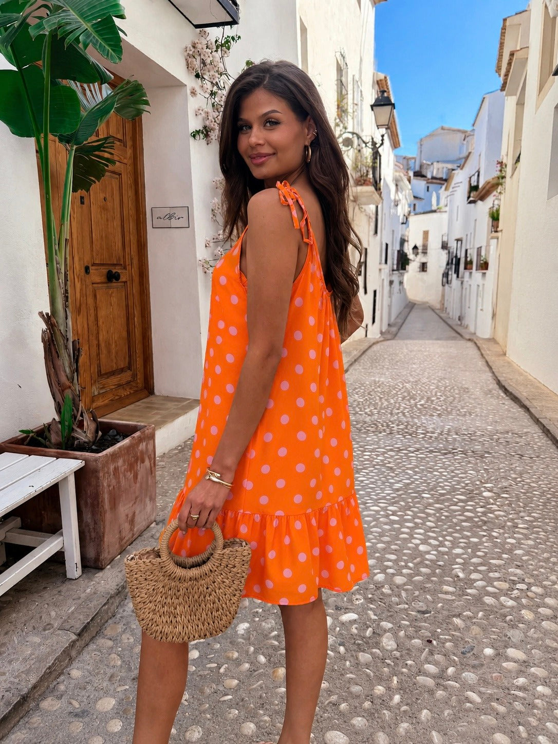 A woman wearing a bright orange sleeveless dress with white polka dots stands on a cobblestone street in a Mediterranean-style village, holding a woven straw handbag.
