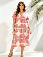 A woman modeling a short-sleeve midi dress with a pink and white citrus slice pattern, standing against a white wall with palm leaf shadows.