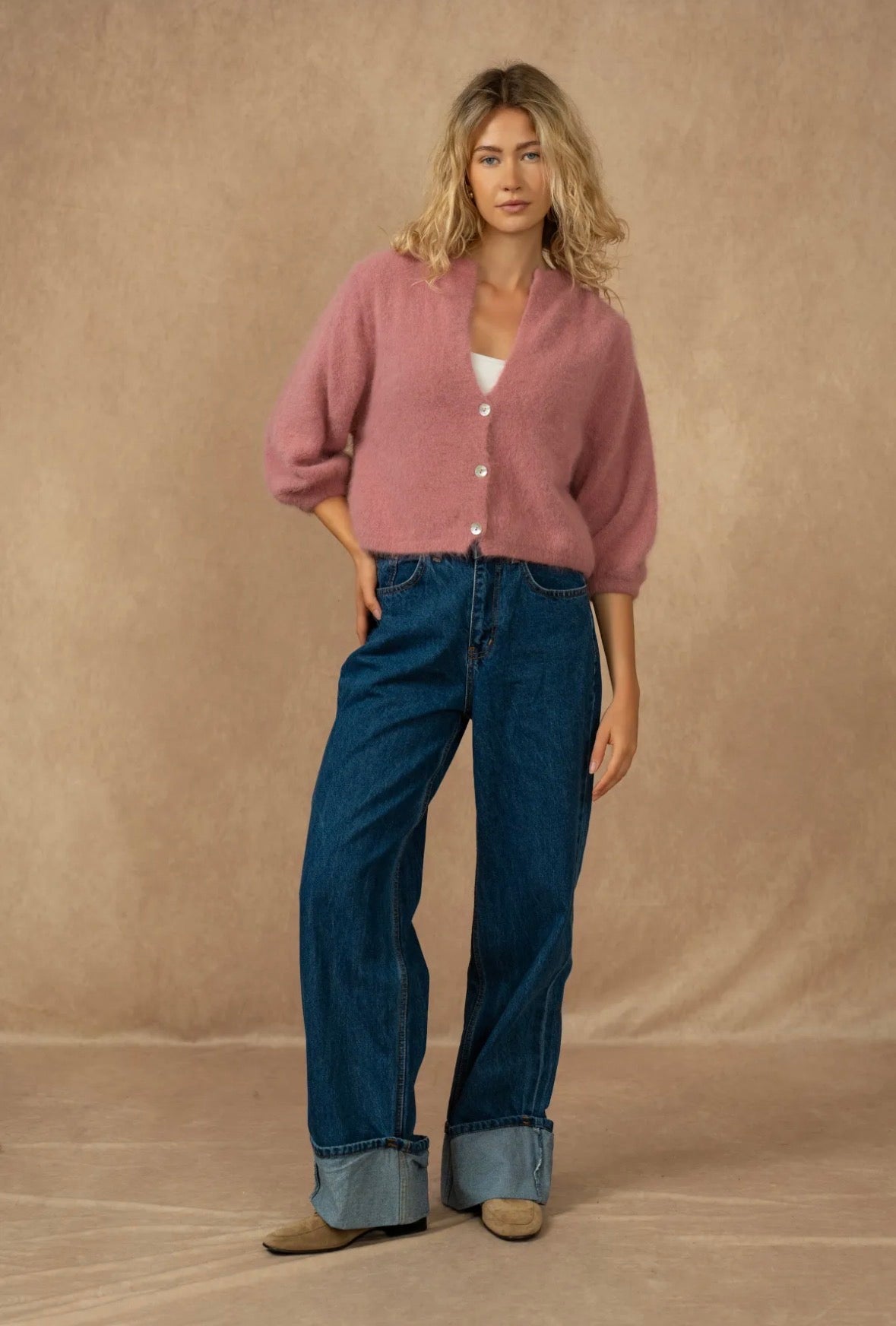 A woman modeling a dusty pink button-up cardigan with three-quarter sleeves, paired with dark blue wide-leg jeans and tan shoes, set against a warm beige studio backdrop.