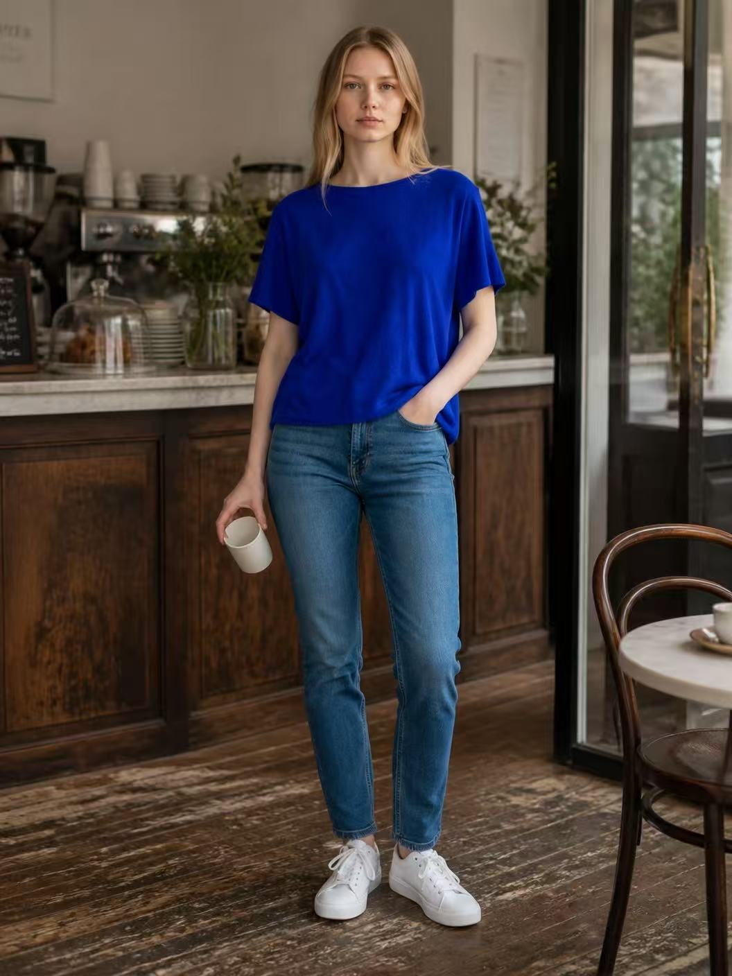 A woman stands in a cafe wearing a vibrant royal blue short-sleeve top, blue jeans, and white sneakers, holding a white mug.