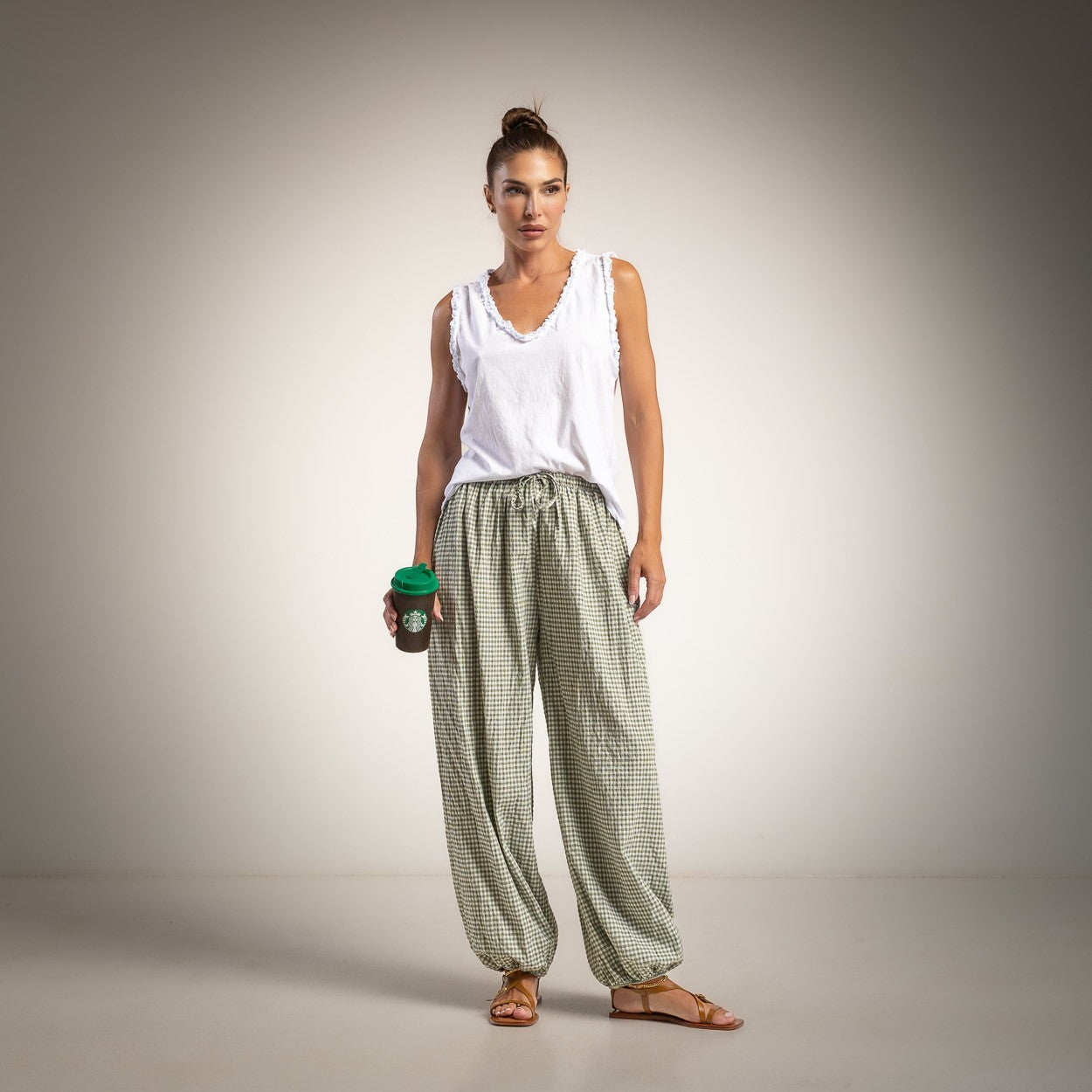 A woman stands against a neutral studio background wearing a white sleeveless top and loose green-and-white checkered pants, holding a dark coffee cup with a green lid.
