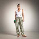 A woman stands against a neutral studio background wearing a white sleeveless top and loose green-and-white checkered pants, holding a dark coffee cup with a green lid.