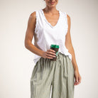 A woman wearing a white sleeveless top with ruffled trim and green-and-white checkered wide-leg pants, holding a dark coffee cup with a green lid against a plain light beige studio background.