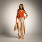 A woman stands against a neutral studio backdrop wearing a bright orange sleeveless top and beige-and-brown vertically striped harem-style pants, holding a tan tote bag.