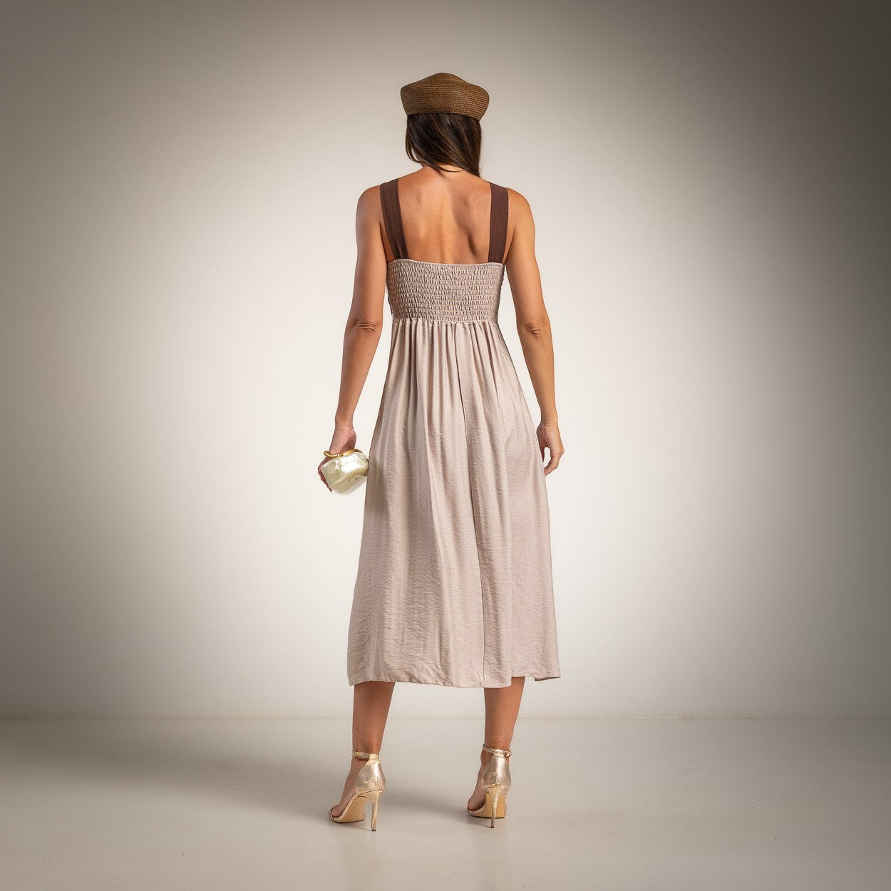 A woman is shown from behind wearing a light taupe midi dress with brown shoulder straps and a shirred back