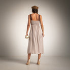 A woman is shown from behind wearing a light taupe midi dress with brown shoulder straps and a shirred back