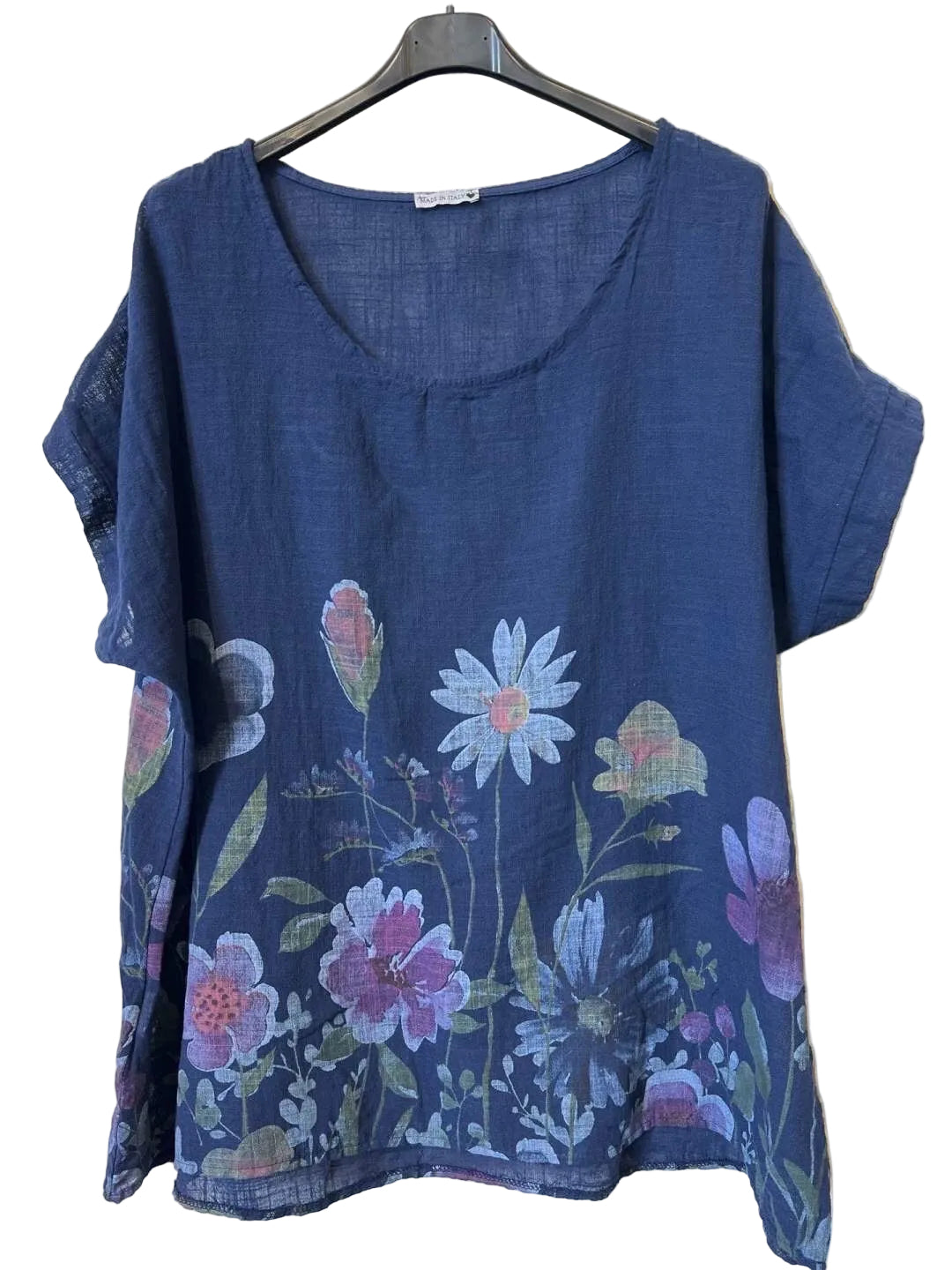 A short-sleeve blue top with a colorful floral print, hanging on a black hanger against a textured light gray wall.