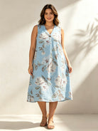 A woman modeling a sleeveless light blue midi dress with a white and gray floral print, standing against a warm beige wall with soft shadows.