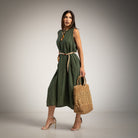 A woman models a sleeveless khaki green midi dress with a beige tie belt, holding a woven straw tote bag