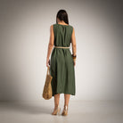 A woman stands with her back to the camera wearing a sleeveless olive green midi dress with a thin beige belt