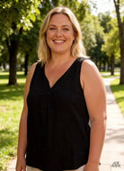 Woman wearing a black sleeveless top standing in a park with trees and grass in the background.
