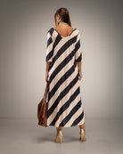 A woman viewed from the back wearing a loose, knee-length dress with bold diagonal black and cream stripes. She is holding a brown leather tote bag