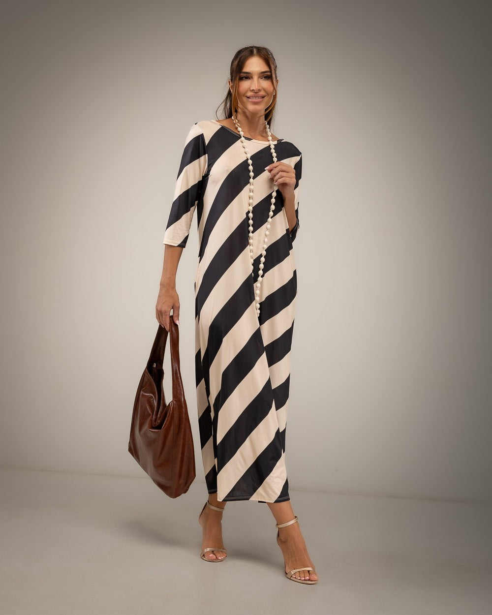 A woman modeling a black and cream diagonal striped midi dress with elbow-length sleeves, holding a brown leather tote bag 