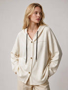 A woman wearing an oversized cream-colored hooded cardigan with black drawstrings and gold-tone buttons, paired with matching cream pants, photographed against a neutral light gray studio background.