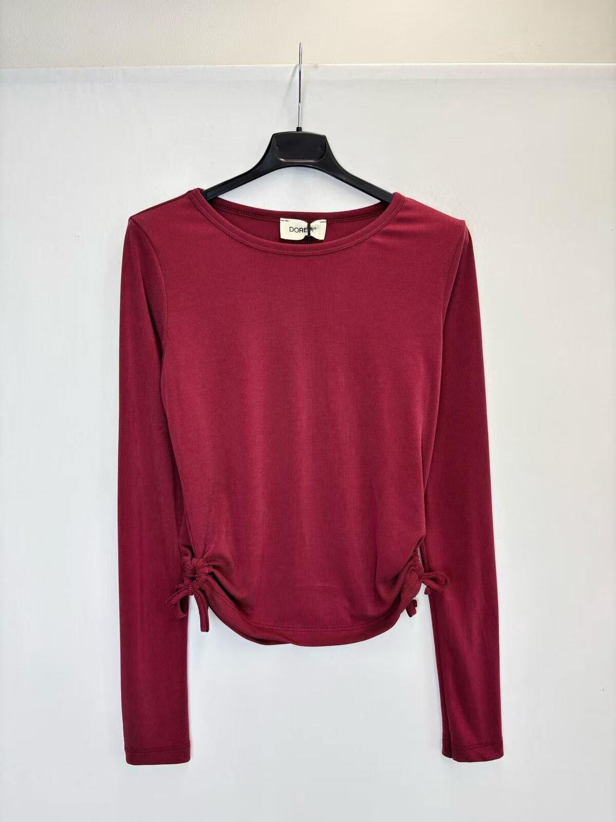 A long-sleeve crewneck top in deep burgundy, hanging on a black hanger against a plain white wall. 