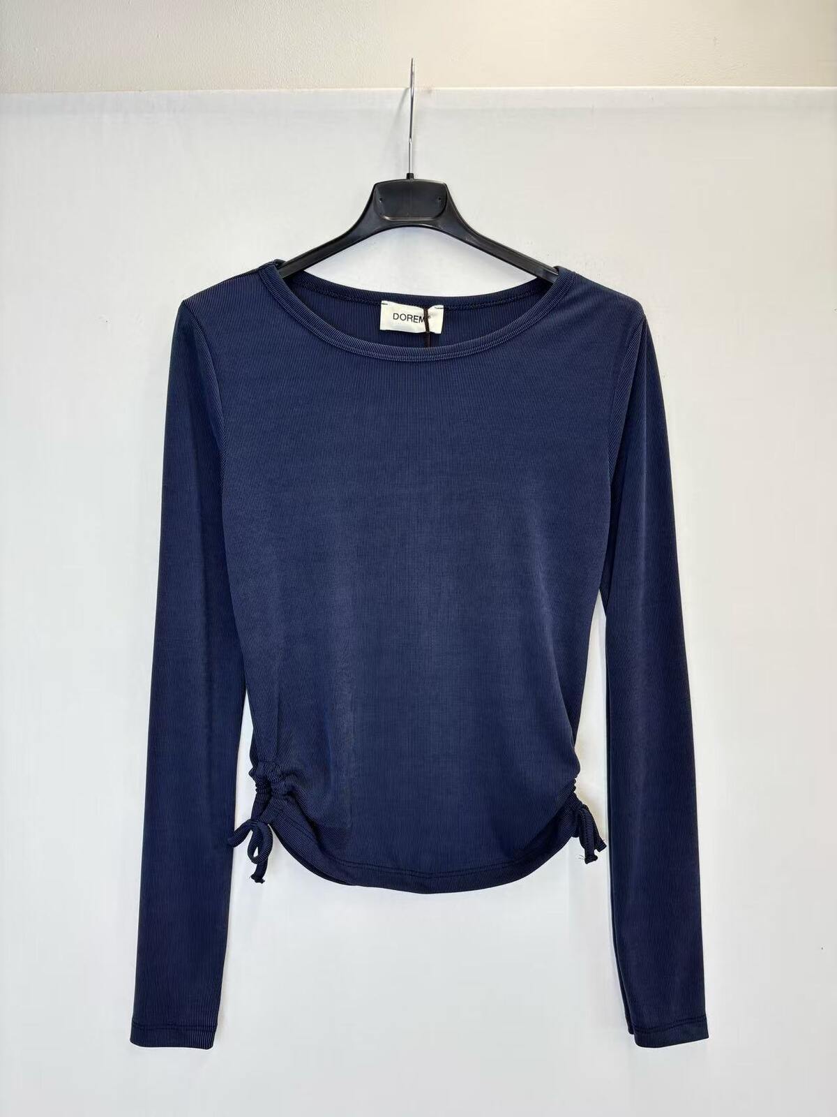 A navy blue long-sleeve knit top hanging on a black hanger against a plain white wall. The top has a crew neckline and side tie details at the hem.