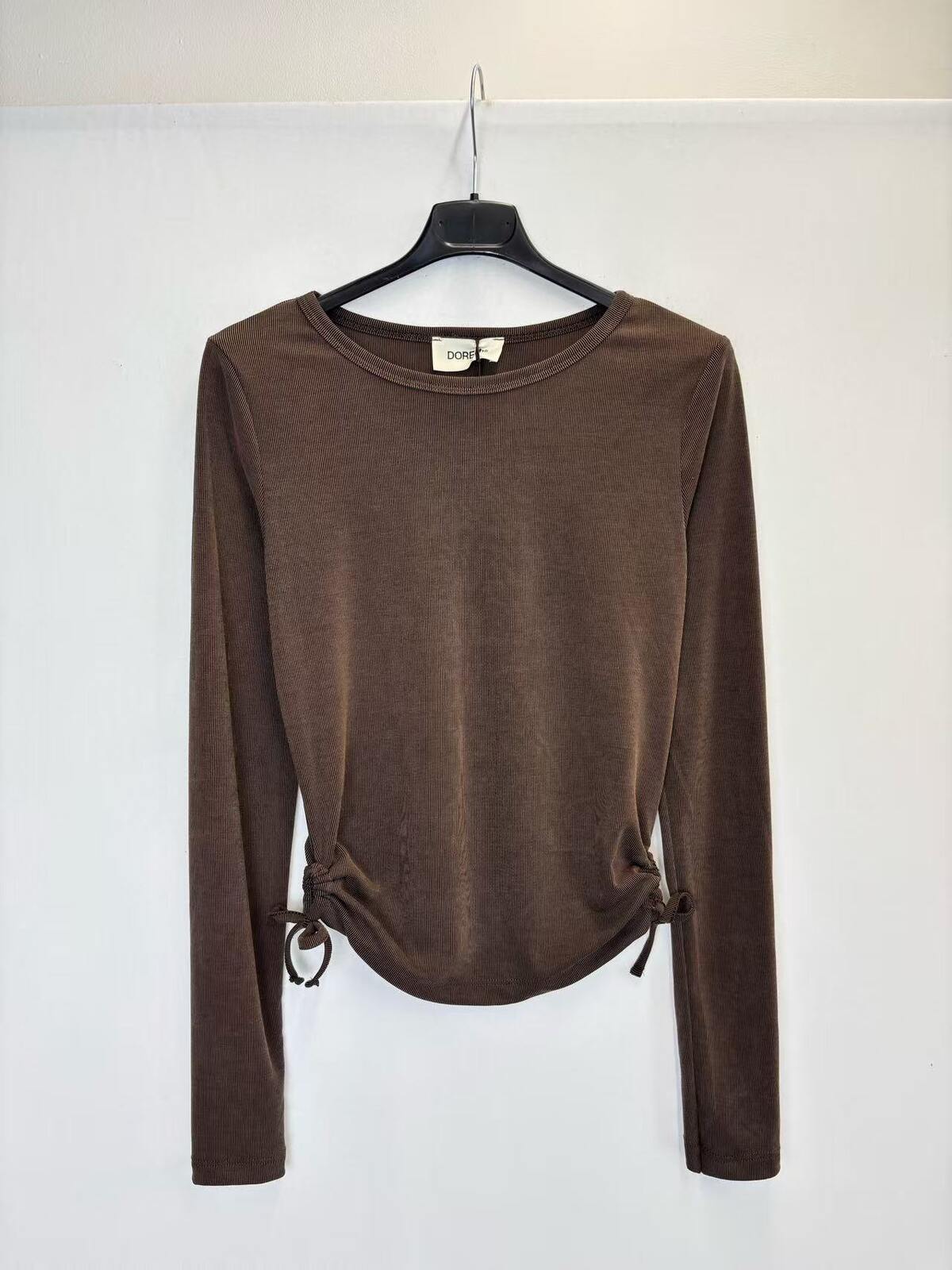 A brown long-sleeve knit top hanging on a black hanger against a plain white wall. The top has a crew neckline and side drawstring details at the hem.