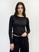 A woman modeling a black long-sleeve crewneck top with side drawstrings, paired with black drawstring pants