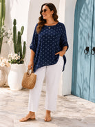 Woman in a blue polka dot top and white pants standing in front of a white wall with a teal door.