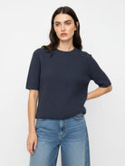 A woman modeling a dark navy blue ribbed short-sleeve knit top with light blue jeans, standing against a plain white studio background.