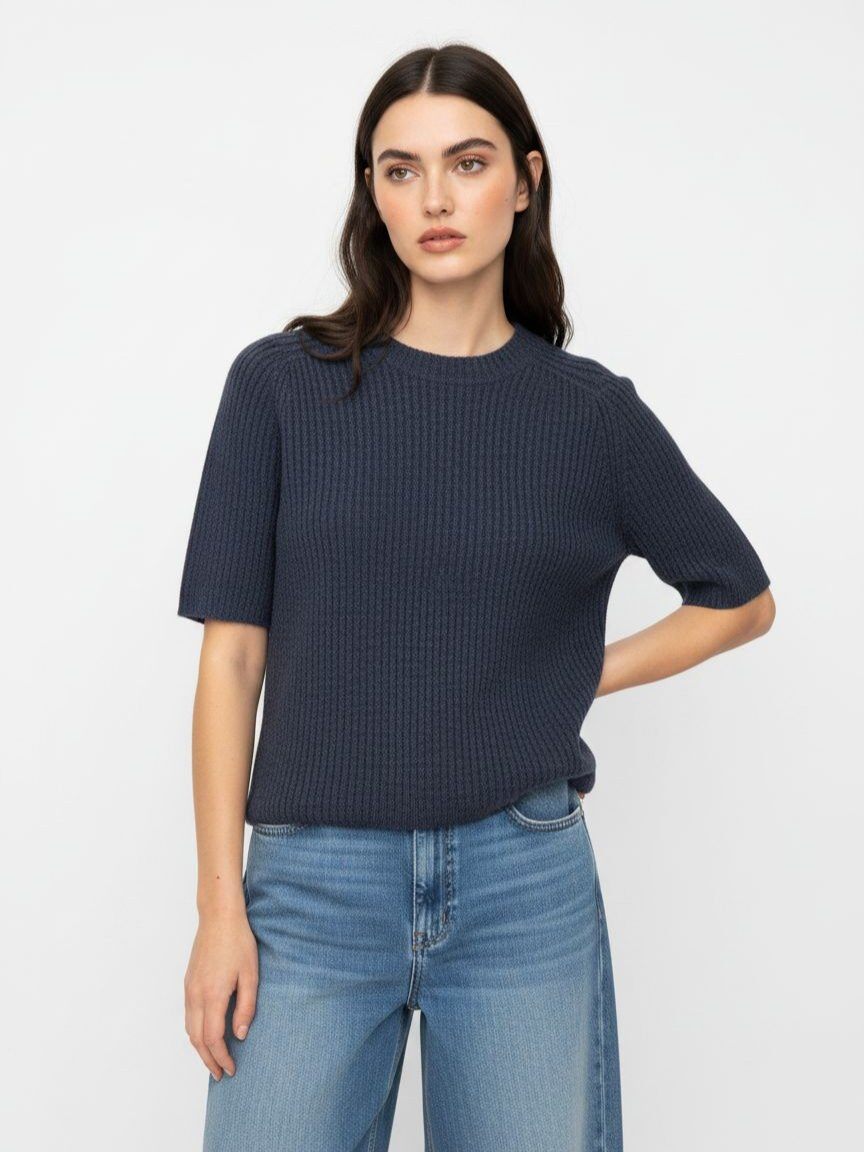 A woman modeling a dark navy blue ribbed short-sleeve knit top with light blue jeans, standing against a plain white studio background.