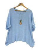 A light blue short-sleeve top with a relaxed, slightly oversized fit, hanging on a wooden hanger against a textured light gray fabric backdrop. A wooden pendant necklace is draped over the front of the shirt.