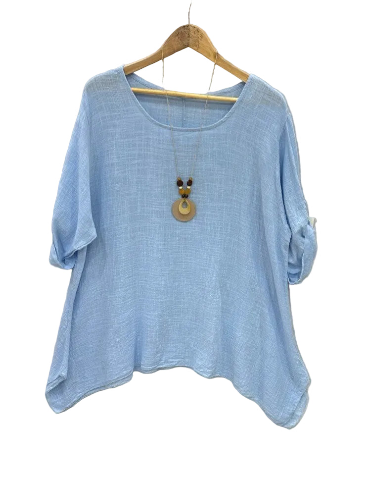 A light blue short-sleeve top with a relaxed, slightly oversized fit, hanging on a wooden hanger against a textured light gray fabric backdrop. A wooden pendant necklace is draped over the front of the shirt.
