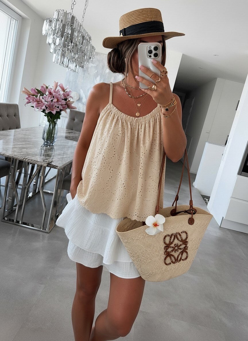 A woman takes a mirror selfie in a modern, bright room, wearing a beige eyelet camisole top, a white ruffled skirt, a straw hat, and carrying a beige woven tote bag with a white flower accent.