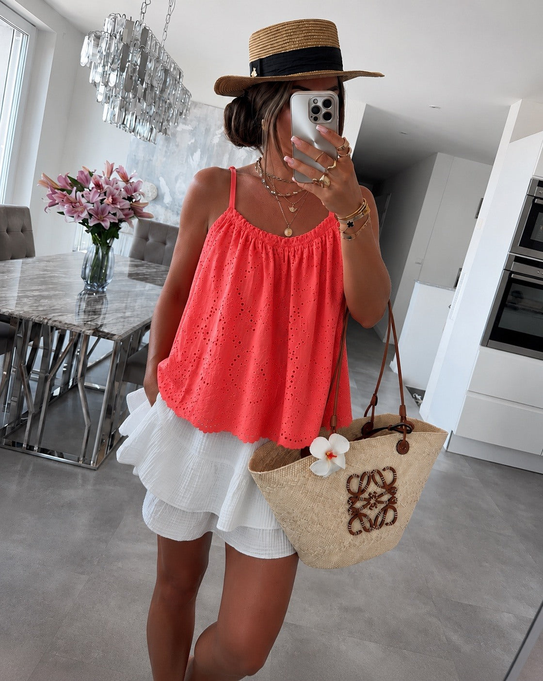 A woman takes a mirror selfie in a modern dining room, wearing a bright coral pink eyelet spaghetti-strap top, a white ruffled mini skirt, a straw hat with a black band, and holding a beige woven straw tote bag with a white flower accent.