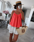 A woman takes a mirror selfie in a modern dining room, wearing a bright coral pink eyelet spaghetti-strap top, a white ruffled mini skirt, a straw hat with a black band, and holding a beige woven straw tote bag with a white flower accent.