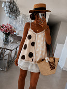 A woman takes a mirror selfie wearing a beige camisole top with large black and white polka dots, white ruffled shorts, a straw hat, and carrying a woven straw tote bag in a modern indoor setting.