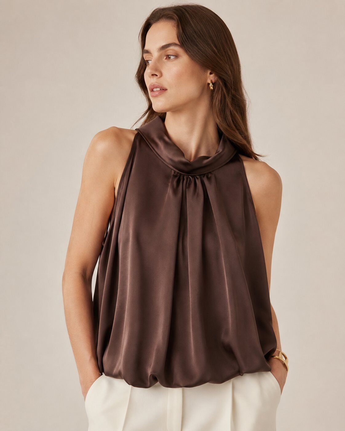 A woman wearing a sleeveless high-neck satin top in dark brown, paired with white pants, photographed against a plain beige studio background.