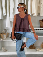 A woman posing in a clothing store wearing a sleeveless mauve-purple ruffled top, light-wash blue jeans, and white sunglasses, holding a black quilted handbag.