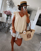 A woman wearing a cream sleeveless V-neck top, white ruffled shorts, a straw hat with a black band, and carrying a beige woven tote bag 